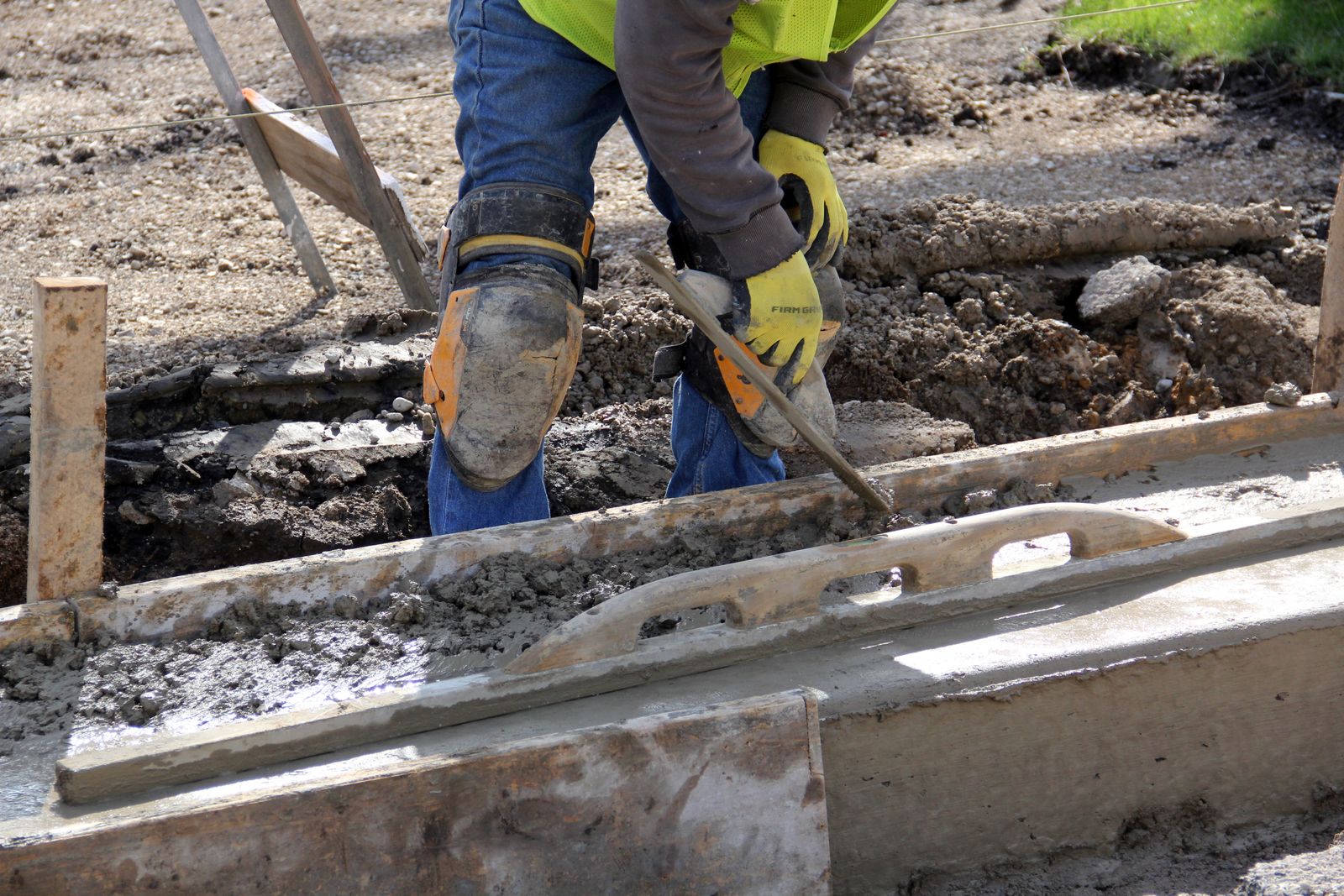 Worker in knee pads and a hi-vis vest setting fresh concrete along wooden curb forms