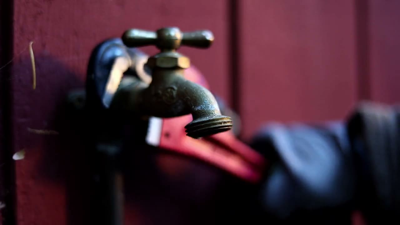 Gloved hand using a red pipe wrench on an outdoor faucet