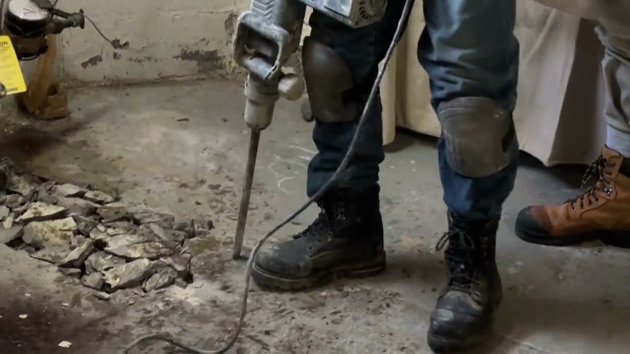 Worker in boots using a jackhammer to break open a concrete floor to reach a drain line